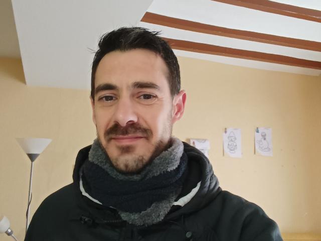 NextLupios ha un sorriso compiaciuto, capelli neri corti e una barba. Indossa una felpa nera con una sciarpa grigia spessa, ed è in piedi all'interno, con luci a soffitto inclinate e opere d'arte alle pareti..