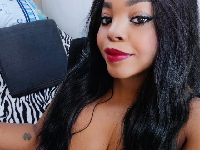 Tatiana Marin, avec de longs cheveux noirs, portant du rouge à lèvres, regardant l'appareil photo avec un léger sourire. Des coussins noirs avec un motif zébré en arrière-plan, et un petit tatouage floral sur son bras supérieur. Extrêmement séduisant..