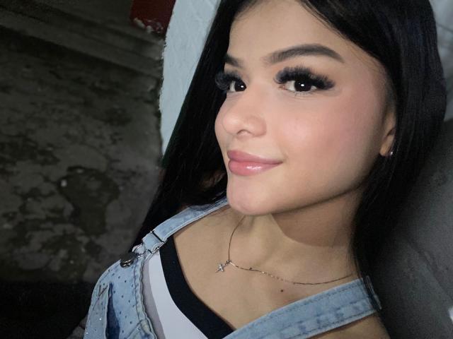 Primo piano di AmberGrayy, una donna attraente con lunghi capelli neri, un trucco occhi marcato e labbra piene, che indossa una camicetta di jeans blu e collana sottile, che guarda direttamente la fotocamera con un leggero sorriso.