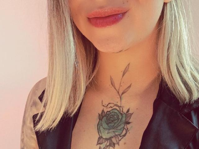 Close-up of KayleeM's neck and chin, showing a green rose tattoo with black outlines. KayleeM has light blonde hair, red lipstick, and is wearing a black leather jacket. Fucking sexy.