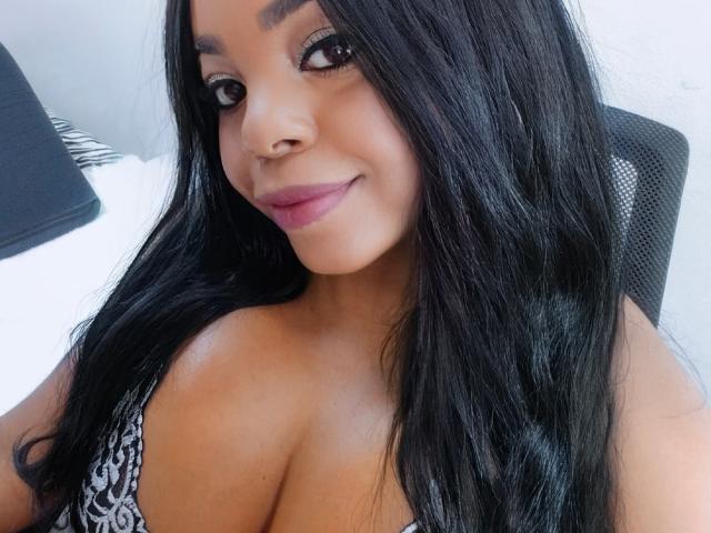 Close-up of TatianaMarin, a curvy woman with long black hair, brown skin, and large breasts, smiling at the camera. She's wearing a low-cut black and white patterned top, exuding sexual tension.