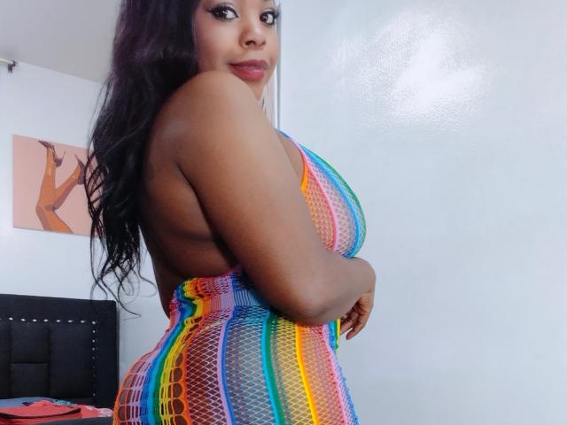 Tatiana Marin, con la pelle di colore marrone scuro, i capelli lunghi e neri, indossa un abito a rete multicolore che mette in risalto le sue forme. seni grandi e glutei rotondi. Si guarda indietro con un'espressione seducente..