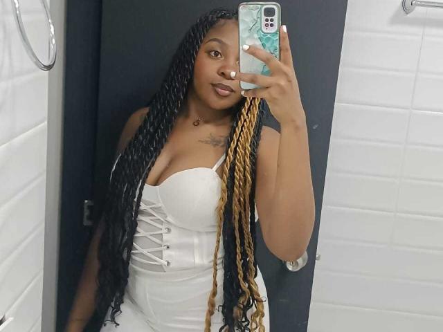 Selfie de Xhaniyah, uma mulher negra com cabelos longos trançados, vestindo um corpete branco justo que realça seus seios grandes. Segurando um telefone com uma capa azul e branca, sorrindo de forma sedutora..
