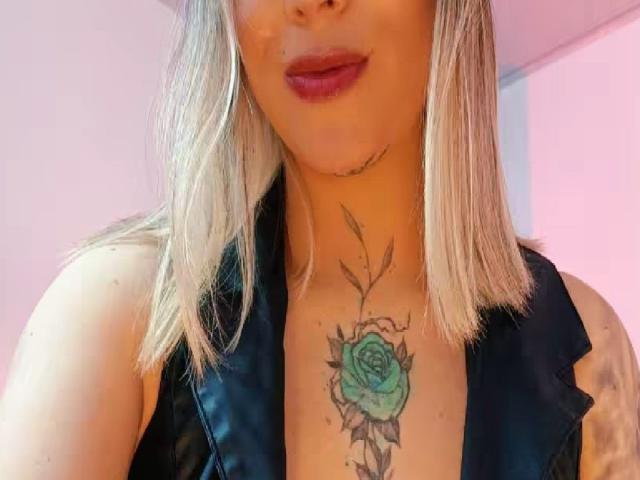 Close-up of KayleeM's neck and upper chest, showing a green rose tattoo with black outlines and thin lines. KayleeM has shoulder-length blonde hair and dark red lipstick. Wearing a black top.