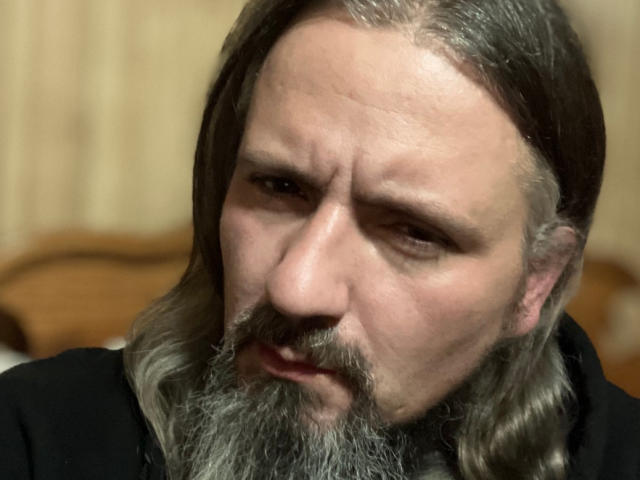 Photo en gros plan de xbillisticX1, avec de longs cheveux et une barbe gris, regardant intensément l'appareil photo. Le front plissé et la bouche légèrement ouverte de xbillisticX1 suggèrent une tension sexuelle. Éclairage doux, arrière-plan flou.