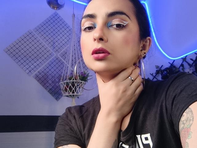 Nahaufnahme von CandyParis mit dunklem Haar, blauem Lidschatten und pinkem Lippenstift, die Hand berührt den Hals, schwarzes Hemd, sichtbar. Tattoo am Arm, Hintergrund mit blauem Neonlicht, hängende Pflanze, Gitterwand..