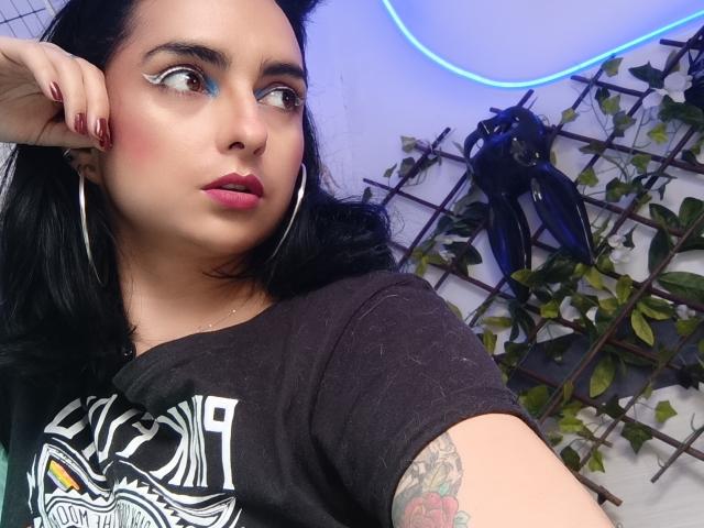 CandyParis, une femme aux formes généreuses, à la peau pâle et aux cheveux noirs, regarde de côté avec du fard à paupières bleu, et porte un t-shirt noir. avec des graffitis blancs. Lumière néon bleue, mur couvert de lierre en arrière-plan. Tatouage sur le bras..