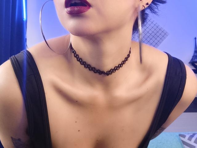 Nahaufnahme von CandyParis mit glänzenden, dunkelroten Lippen, die einen schwarzen Choker und Ohrringe im Hula-Hoop-Stil trägt. Sie trägt ein schwarzes Tanktop, das ihren Ausschnitt und ihren Schlüsselbein betont. Der Hintergrund ist blau und unscharf..