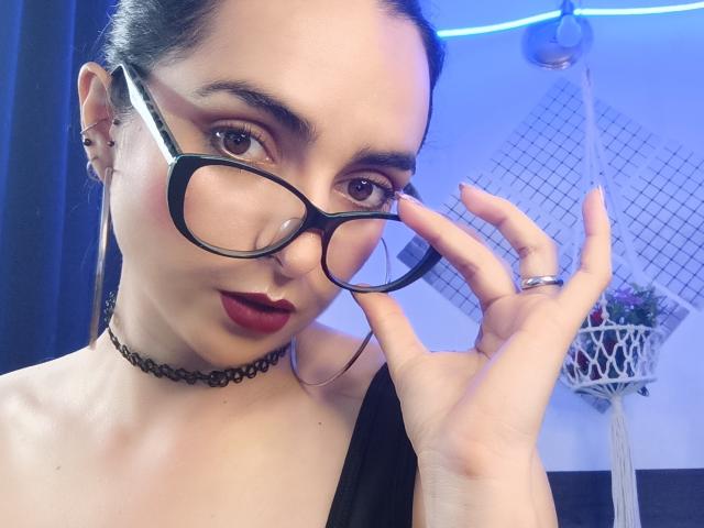 Nahaufnahme von CandyParis, mit heller Haut und dunklem Haar, die eine Brille mit schwarzem Rahmen, roten Lippenstift, eine schwarze Halskette und Schwarz oben. Brille in einer Hand haltend, Hintergrund mit blauem Licht, Makramee-Pflanzenaufhänger. Verdammt heiß..