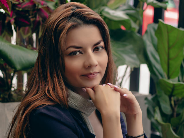 Photograph of MandyMouses, a brunette woman with long hair, brown eyes, and light skin, holding her chin with her right hand. She wears a black jacket with a gray scarf. Green plants in the background. Fucking cute, sexy vibe.