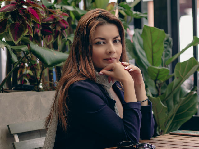Photograph of MandyMouses, sitting at a wooden table, leaning on her chin with a sultry expression. She's wearing a black top, gray scarf, brown hair, surrounded by green plants. Her hand rests on her cheek, sunglasses on the table.