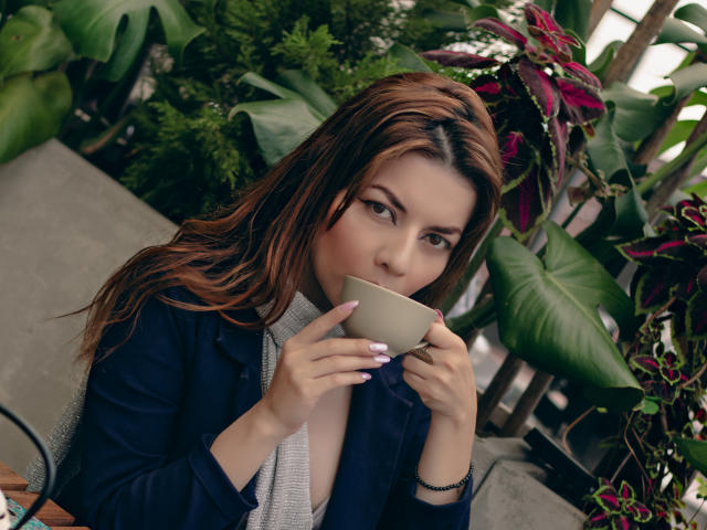 MandyMouses con cabello castaño largo, sosteniendo una taza beige, vistiendo un blazer negro sobre una blusa blanca, rodeadas de exuberante vegetación verde. plantas. Ligera sonrosadura, mirada intensa, tensión sexual.