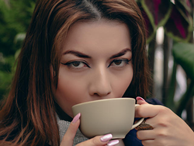 Close-up of MandyMouses, a young woman with brown hair and heavy eyeliner, holding a white cup to her lips. Her intense gaze is focused forward, with green foliage blurred in the background.