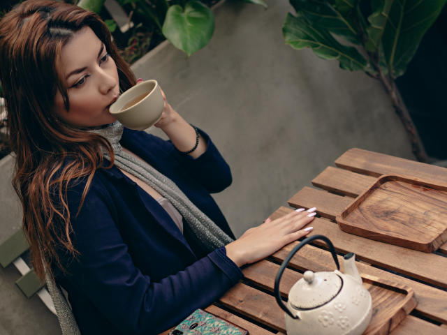 MandyMouses, avec ses cheveux bruns, sirotant une boisson dans une tasse blanche, porte une veste noire et une écharpe grise, et se penche sur une table en bois. théière blanche, feuilles vertes en arrière-plan, ambiance détendue et séduisante..