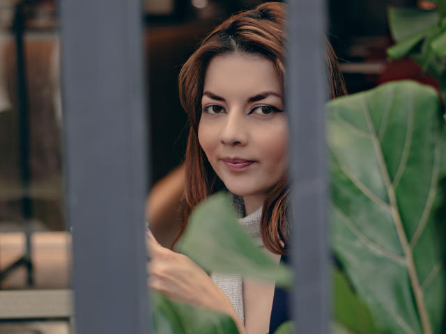 Foto van MandyMouses, een bruinharige vrouw met haar tot op de schouders, die verleidelijk door groene bladeren heen kijkt, gedeeltelijk zichtbaar. haar borst, zwarte top, subtiele glimlach, zacht licht, onscherpe voorgrond, intieme sfeer, sexy uitstraling.