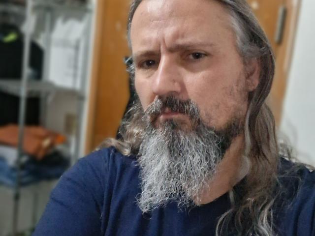 Close-up photo of an older man with a long gray beard and mustache, wearing a black shirt. He looks serious, with light brown eyes and gray hair. Blurred background.