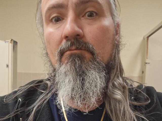 Primer plano de xbillisticX1, un hombre de mediana edad con una barba y bigote largos y grises, mirando directamente a la cámara con una expresión ligeramente sorprendida, vistiendo una camisa negra. Puerta visible en el fondo.