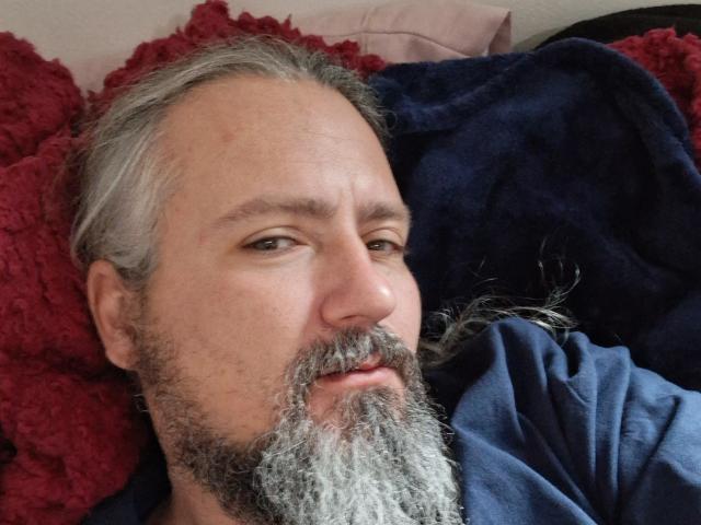 Nahaufnahme eines Fotos von xbillisticX1, einem Mann mittleren Alters mit grauem Bart und Schnurrbart, der auf einer rot-schwarzen, strukturierten Oberfläche liegt. Kissen. Sein Ausdruck ist leicht verführerisch, mit einem Hauch von Überheblichkeit..