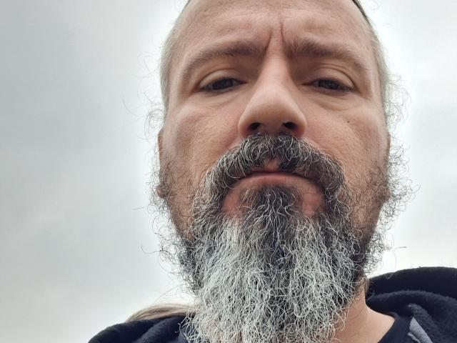 Close-up foto van een kale, middelbare man met een dikke, grijze baard en snor, die naar beneden kijkt met een lichtelijk zelfvoldane blik. uitdrukking.
zijn gezichtsbeharing is opvallend en heeft een duidelijke textuur. Een ongelooflijk intense blik..