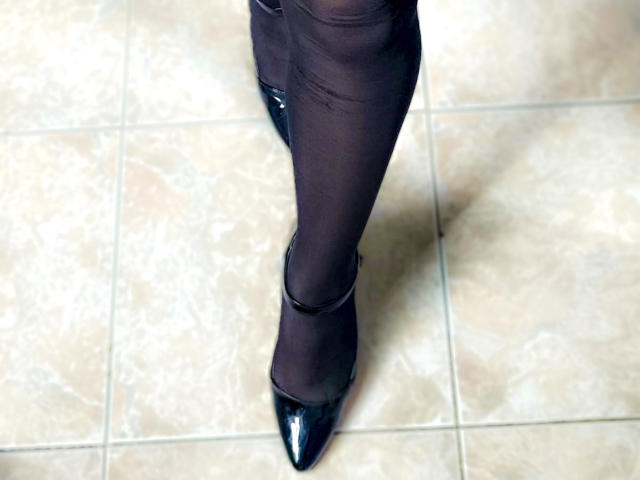 Close-up of MiaBellucci's right leg and foot, wearing black sheer pantyhose and shiny black patent leather pumps, standing on white tiled floor. Slight shadow under foot, highlighting glossy shoe.