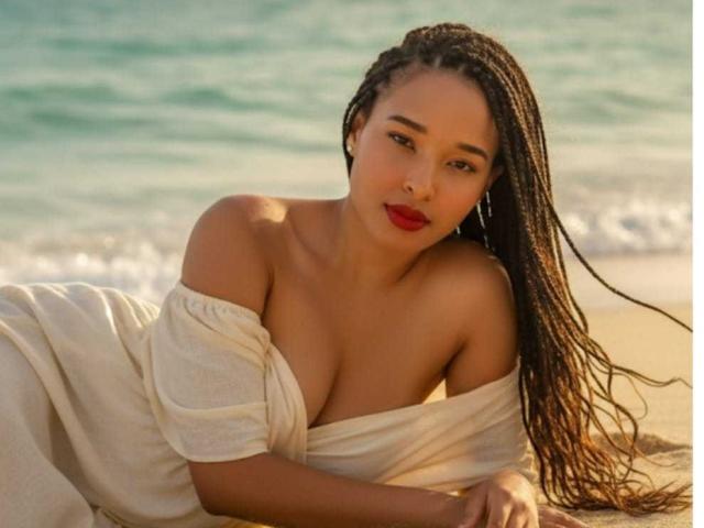 AmelyGomes mit langem, nassem, schwarzem Haar, liegt am Strand, trägt ein weißes Kleid mit einem frechen Ausschnitt, das ihre Brust betont, sexy. verführerischer Blick, roter Lippenstift, Ozean im Hintergrund, Sonnenlicht auf der Haut.