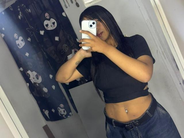 NahomyNoir taking a mirror selfie with an iPhone, wearing a black crop top and dark jeans, showing a pierced navel. Long black hair, slight lean, phone obscures face. Dark curtains with ghost patterns in background.