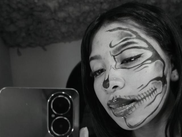 Black and white selfie of NahomyNoir with face painted like a sugar skull, holding a phone with dual cameras. She has shiny lips, dark eye makeup, and a grinning expression.