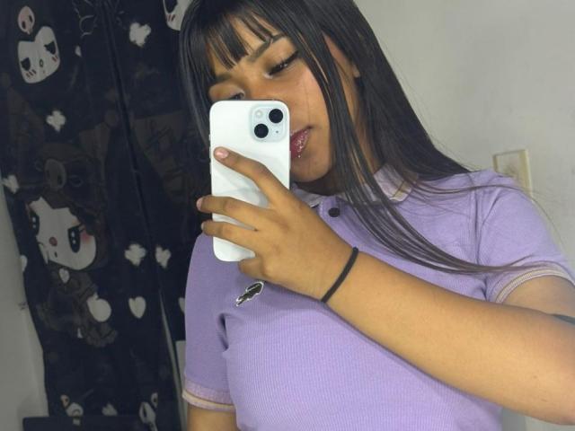 NahomyNoir, a curvy woman with long black hair and bangs, takes a mirror selfie with a white iPhone. She wears a tight purple shirt accentuating her large breasts. Flirty, seductive expression.