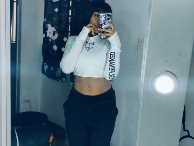 NahomyNoir, selfie, mirror, holding phone, white crop top, "MIND" text, black pants, navel piercing, dark hair, flash reflection, room, dark patterned curtain, casual, sexy, confident, slightly blurred, modern style.