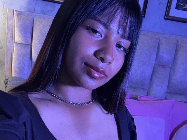 Close-up of NahomyNoir, an Asian woman with shiny, glossy lips and dark straight hair, wearing a black top and silver chain necklace, under blue lighting. Her eyes are half-closed seductively.