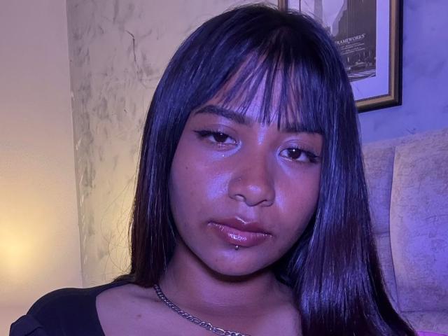 Close-up of NahomyNoir, a dark-skinned woman with straight black hair and bangs, glossy lips, and a chain necklace. Blue lighting highlights her skin, creating a sexy, sultry vibe.