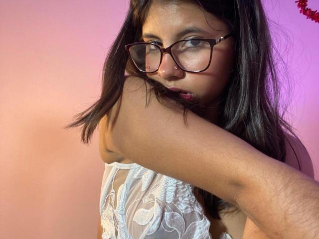 Close-up of CelesteMorel with dark hair, wearing glasses and a white lace top. She's raising her arm, exposing her underarm and gazing intensely. Background has pink and purple gradient.