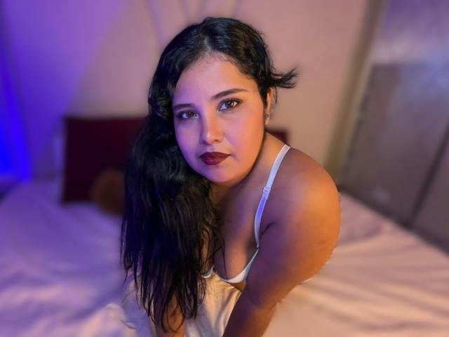 Fotografie von Cassandra Valmont, einer kurvigen Frau mit langen, schwarzen Haaren, die ein weißes Tanktop trägt und auf einem Bett nach vorne gebeugt ist. Ihr Ausdruck ist sinnlich, und die blaue und orangefarbene Beleuchtung betont ihre Haut..