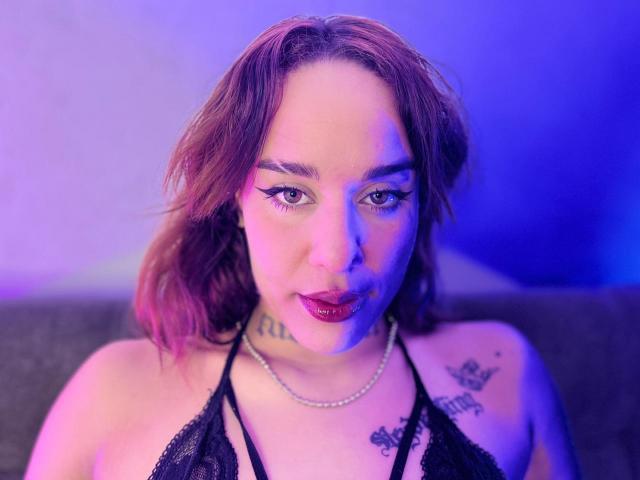 Close-up foto van HappyAndDebora, een vrouw met bruin haar dat tot op haar schouders reikt, met blauwe en paarse verlichting, die een zwart topje met halter, een ketting en tatoeages op haar borst en schouders. Geconcentreerde uitdrukking, lichtjes open lippen.