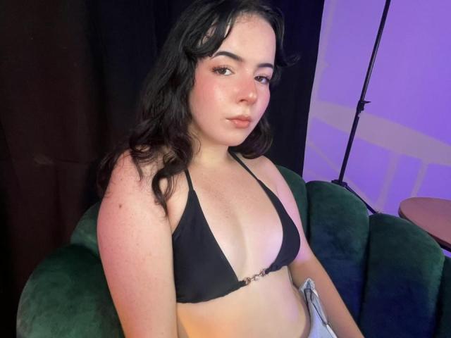 Photograph of AliinaFontaine with pale skin and dark wavy hair, wearing a black triangle bikini top, sitting on a green couch, blue-purple lighting, sexy gaze, slight smile, relaxed pose.