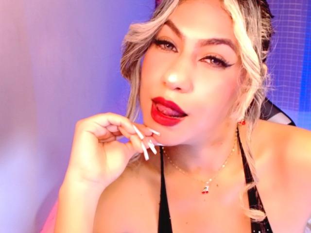 Fotografia ravvicinata di AnastaciaMonaco, capelli biondi, rossetto rosso, top nero con spalline, dito vicino alla bocca, sguardo seducente. illuminazione intensa, sfondo blu, pelle chiara, unghie lucide, sexy, espressione intensa..