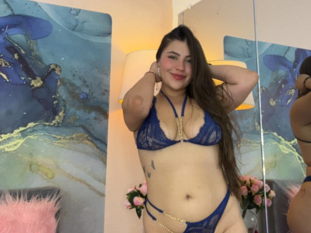 LorensDouce stands in front of a mirror, wearing a blue lace bra and thong, with long brown hair, smiling, arms raised, showcasing her curvy physique and a gold chain across her chest.