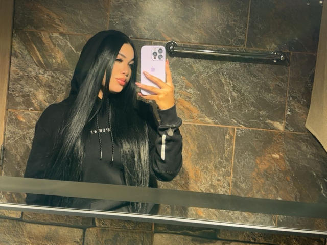 MiaDenver, long black hair, taking a mirror selfie with a white iPhone, wearing a black hoodie, brown tiled wall background, holding phone in right hand, reflective mirror, casual pose.