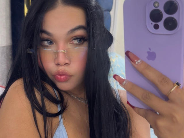 Close-up of MiaDenver, a young woman with long black hair, wearing glasses and a blue tank top, holding a purple iPhone with gold camera lenses. Her nails are long and red. Fucking sexy.