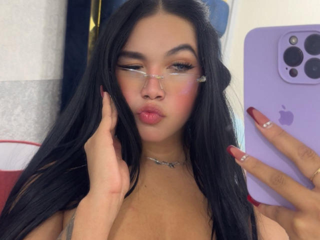 Close-up van MiaDenver die een selfie maakt. Ze heeft lang zwart haar, draagt een bril en heeft roze blush op. Haar hand raakt haar gezicht aan, waarbij een zilveren ketting zichtbaar is. Ze houdt een paarse iPhone vast. Heel erg aantrekkelijk..