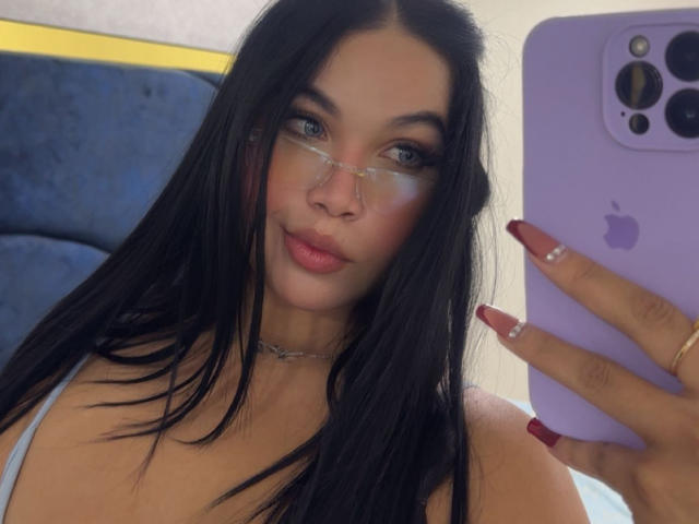 MiaDenver maakt een selfie met een paarse iPhone, die ze in haar rechterhand vasthoudt. Ze heeft lang zwart haar, draagt een bril, heeft rode nagellak en een zilveren ketting. Heel aantrekkelijke uitstraling.