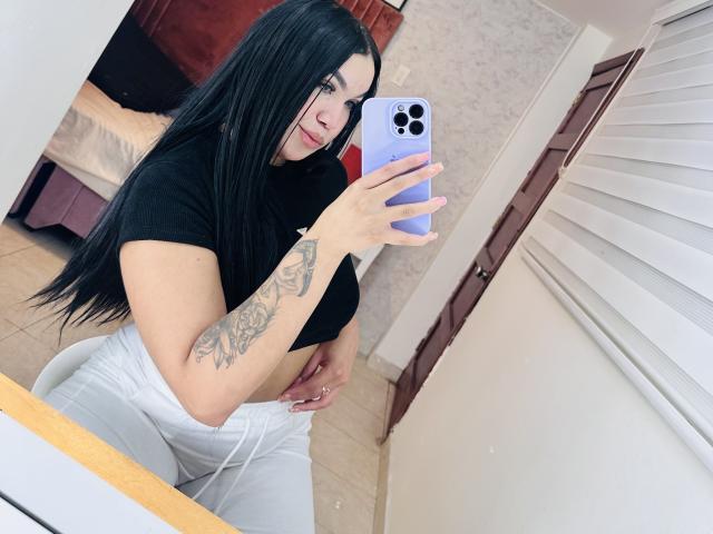MiaDenver with long black hair, holding a white iPhone, taking a mirror selfie. She's wearing a black t-shirt and white pants, showing a tattoo on her right arm. Fucking sexy.
