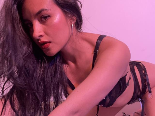 Close-up van EmilyJensenn, voorovergebogen met lang zwart haar, gekleed in zwarte kant lingerie, glanzende huid, rood. lipsticks, oorringen. Het roze licht accentueert haar rondingen, waardoor er een seksuele spanning ontstaat..