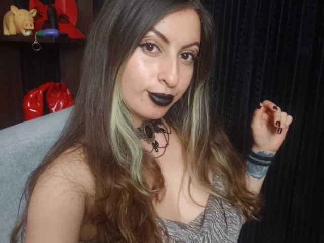 LeilaGoddes tiene el cabello largo y castaño con reflejos verdes, lápiz labial negro y maquillaje de ojos oscuro. Ella lleva una blusa gris de encaje, un collar negro y una pulsera. Se ven cortinas negras al fondo. Increíblemente atractivo/a..