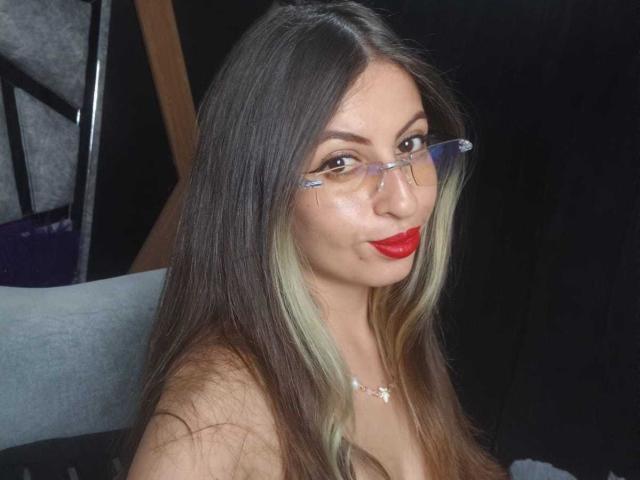LeilaGoddes, con cabello largo y negro con reflejos rubios, usa gafas y tiene pintalabios rojo, y tiene una expresión seductora. Está sin sujetador, con el escote visible, sobre un fondo oscuro. Increíblemente atractivo/a..