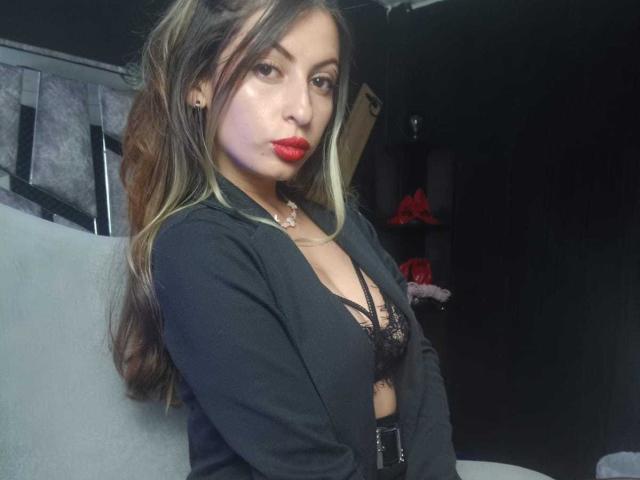 LeilaGoddes, een Aziatische vrouw met lang, golvend zwart haar en rode lippenstift, draagt een zwarte cardigan over een zwarte kanten BH. Zittend op een grijze bank, donkere achtergrond, intense blik, sensuele uitstraling..