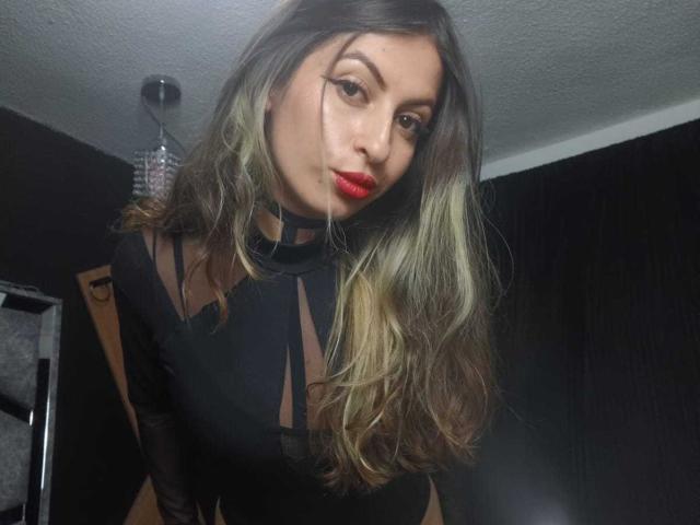 LeilaGoddes has long, wavy brown hair with blonde highlights, wearing a black sheer top with a choker. She's leaning forward, red lips, sultry gaze, dark room, dim light. Fucking sexy.
