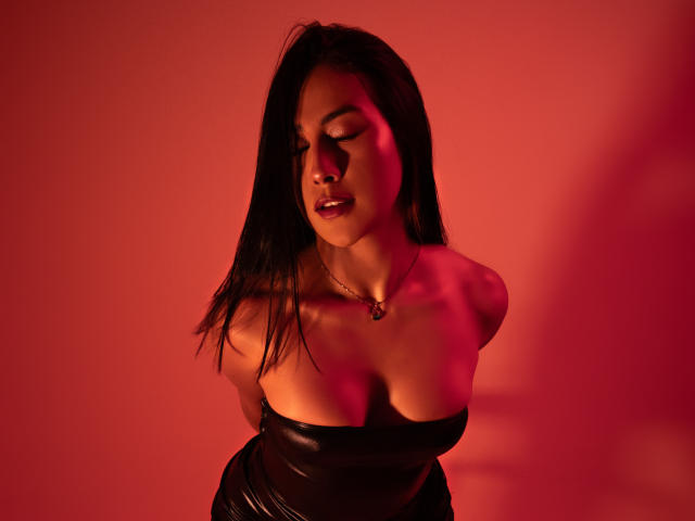Photograph of AnyaJay, a sexy woman with long black hair, leaning forward with eyes closed, wearing a black strapless top, red light casting dramatic shadows, emphasizing her cleavage and sultry expression.