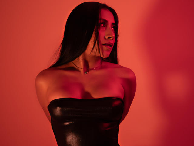 Fotografia de AnyaJay com cabelos longos e pretos, fundo iluminado em vermelho, vestindo uma blusa de couro preta sem alças, olhando para... à direita, sombras que realçam seu busto, iluminação vermelha intensa que cria tensão sexual..