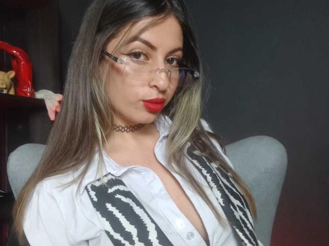 Photograph of LeilaGoddes, Asian woman with long dark hair, red lipstick, and glasses, wearing an open white shirt with black stripes, exposing cleavage. Choker necklace, red object in background. Sexual tension.